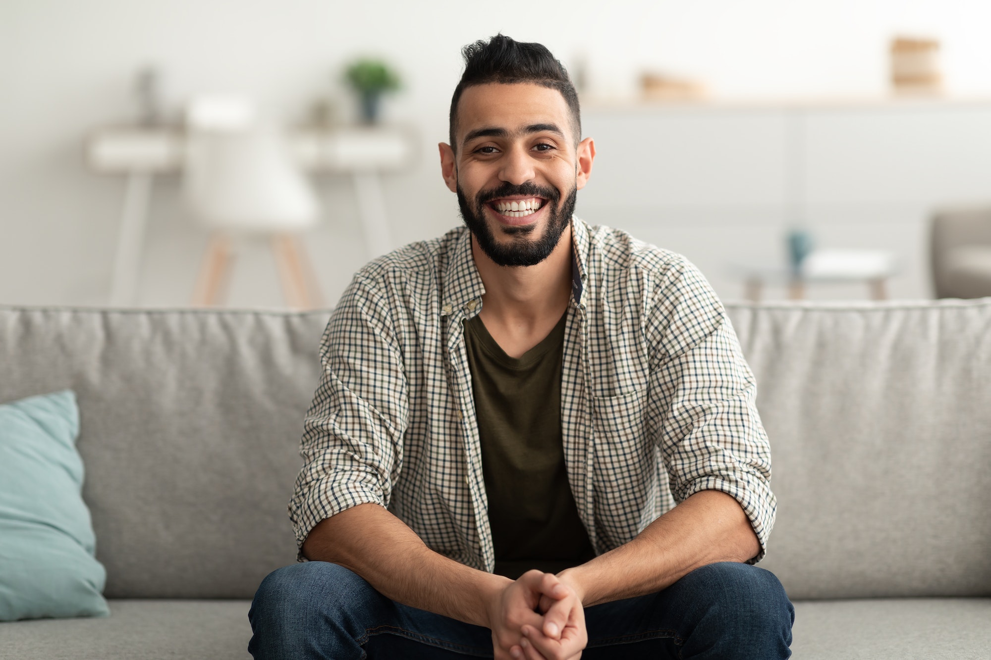 Portrait of handsome young Arab man smiling and looking at camera, sitting on sofa at home